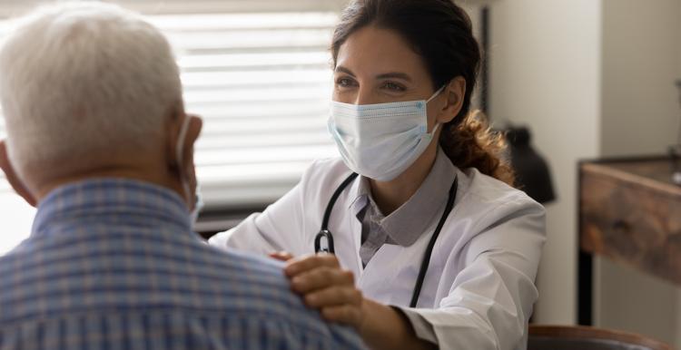 Health professional in a white coat and mask engaging with an elderly man wearing a check shirt.