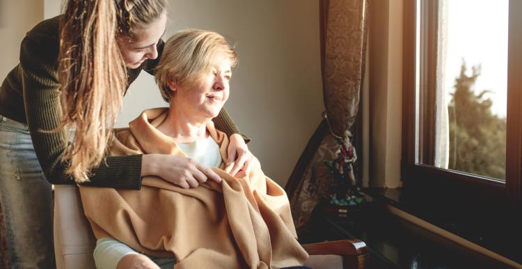 Young woman wraps a blanket around a middle-aged woman sitting in a chair looking out the window