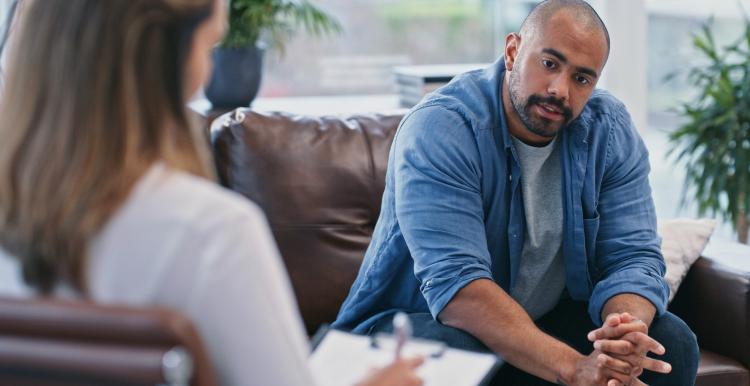 Black man sitting on a sofa talking to a woman who is holding a clipboard