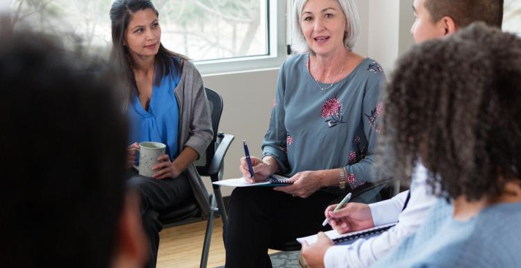 White-haired lady takes notes in a meeting with a group of diverse people sitting in a circle.