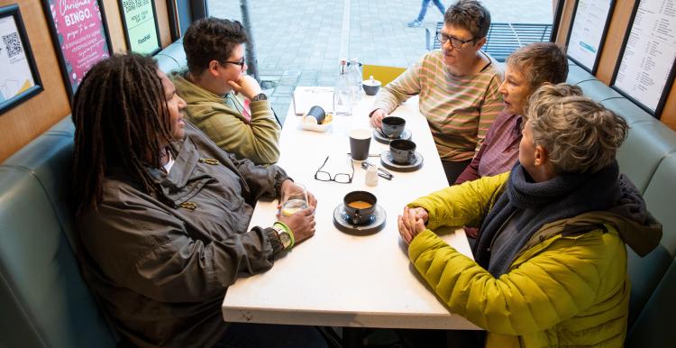Group of five adults sitting at a table near a window drinking coffee and talking