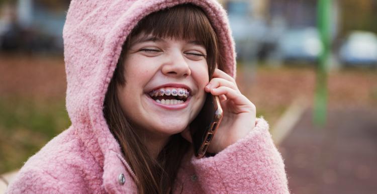 Young girl with braces in a pink hooded jacket laughing and talking on the phone