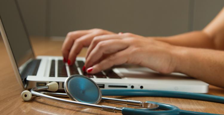 Close-up image of hands typing on a laptop keyboard with a stethoscope lying on the desk