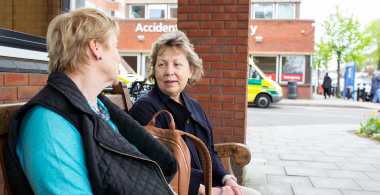 Two women sitting on a bench outside a hospital with ambulance in background