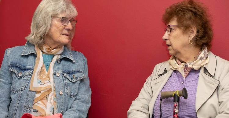 Two older ladies sitting and talking with a red background behind