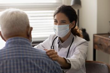 Health professional in a white coat and mask engaging with an elderly man wearing a check shirt.