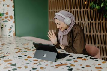 a woman sitting at a table smiling and waving at a laptop