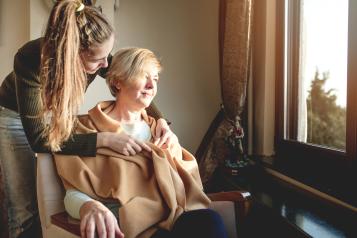 Young woman wraps a blanket around a middle-aged woman sitting in a chair looking out the window