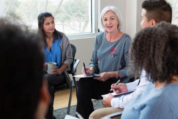 White-haired lady takes notes in a meeting with a group of diverse people sitting in a circle.