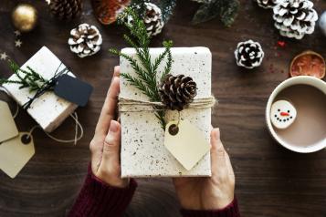 Hands holding wrapped present with pine leaves and a pinecone and hot drink next to it