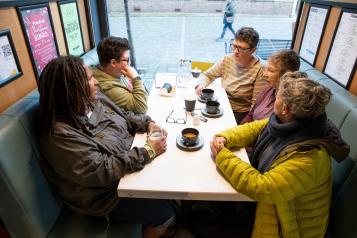 Group of five adults sitting at a table near a window drinking coffee and talking