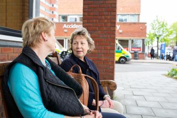 Two women sitting on a bench outside a hospital with ambulance in background