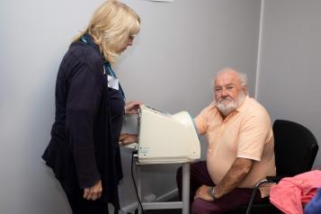 Older white man having his blood pressure checked with a lady standing alongside to help.