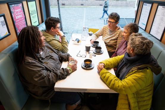Group of five adults sitting at a table near a window drinking coffee and talking