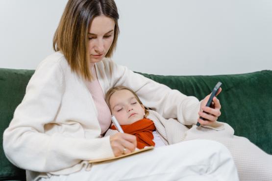 White lady holding on the phone, taking notes, sitting with her sick child.