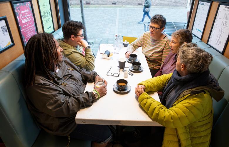 Group of five adults sitting at a table near a window drinking coffee and talking