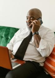 Black man sitting on a green sofa talking on the phone while looking at a laptop